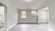 This single-floor unit offers a clean, modern aesthetic achieved through a thoughtful renovation. The living area is generously sized, featuring light gray walls, crown molding, white baseboards, and light hardwood-style flooring that extends throughout the main spaces. Multiple windows fitted with wood-toned blinds allow natural light to fill the room, and a wall-mounted air conditioning unit provides climate control. An elegant arched doorway connects the living area to adjacent spaces, adding a subtle architectural detail to the otherwise understated design.

The kitchen has been fully updated with white shaker-style cabinetry accented by matte black hardware, white quartz countertops, and a classic white subway tile backsplash. Stainless steel appliances, including a refrigerator, gas range, and over-the-range microwave, complete the functional and contemporary cooking space.

The bathroom is tastefully finished with white subway tile surrounding the bathtub, wood-look tile flooring, and matte black fixtures that add contrast throughout. The bedroom features soft gray carpeting, crown molding, and neutral walls, creating a comfortable and quiet retreat. An entry area with hardwood-style flooring and white panel doors rounds out this well-appointed space.