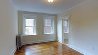 This single-floor residence showcases a classic architectural character evident in its well-maintained hardwood floors, crisp white crown molding, and substantial baseboards throughout. The main room is bright and airy, benefiting from two double-hung windows with traditional white trim that frame views of a neighboring brick exterior. Natural light fills the space, highlighting the warm tones of the hardwood flooring. A third window is visible through a doorway, suggesting an adjoining room that shares the same light-filled quality. The walls are finished in a soft, neutral cream tone that complements the clean white millwork, creating a cohesive and timeless aesthetic. A ceiling-mounted light fixture provides additional illumination. The hallway connecting the rooms is clean and well-proportioned, with matching hardwood floors and consistent white trim detailing that carries the design language throughout the unit. Crown molding lines the ceiling of the corridor, reinforcing the traditional character of the space. The layout appears functional and straightforward, with rooms flowing naturally from the central hallway. Overall, the space presents a classic, well-preserved interior with enduring architectural details.