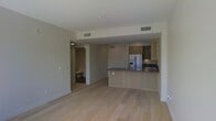 This single-floor residence offers a thoughtfully designed open-concept layout that seamlessly connects the living and kitchen areas. The space is finished with light-toned hardwood flooring that runs throughout, lending a warm and cohesive feel to the interior.

The kitchen is a standout feature, equipped with stainless steel appliances including a French door refrigerator and a built-in oven. Cabinetry in a neutral wood tone lines the walls, complemented by dark countertops that provide a striking contrast. A peninsula with a matching dark countertop extends into the living area, offering additional prep space and casual seating, making it ideal for both cooking and entertaining.

The open living area is bright and airy, with clean white walls and recessed lighting that contribute to a modern, minimalist aesthetic. HVAC vents are integrated seamlessly into the ceiling, ensuring comfort throughout the space.

A hallway visible from the main living area leads to additional rooms, including what appears to be a bathroom with classic fixtures. The overall ambiance is contemporary and understated, with a neutral color palette that provides a versatile backdrop for a variety of interior styles.