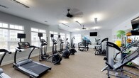 This single-floor fitness center is thoughtfully designed to support a comprehensive workout experience. The space is bright and airy, with rows of large windows along the walls allowing abundant natural light to fill the room throughout the day. The ceiling is finished in crisp white and fitted with dual ceiling fans, ensuring comfortable air circulation during workouts.

The cardio area is prominently featured, housing multiple elliptical trainers and treadmills arranged in an organized row, making efficient use of the open floor plan. The flooring is a neutral carpet, providing a comfortable surface underfoot. A free weights station lines one wall, complete with a full dumbbell rack and kettlebells, catering to a range of strength training needs. An expansive wall mirror reflects the space, enhancing the sense of openness while allowing users to monitor their form.

A wall-mounted flat-screen television is positioned for easy viewing from various points in the room, adding an entertainment element to workout sessions. Additional strength and resistance equipment rounds out the facility, ensuring users have access to a well-rounded range of training options. The clean, neutral color palette and organized layout create a focused, motivating environment suitable for all fitness levels.