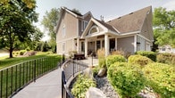 This single-floor home presents an inviting craftsman-style exterior clad in warm taupe siding with crisp white trim accents. A prominent gabled roofline with an elegant arched window crowns the facade, while a covered front porch with sturdy columns provides a welcoming entryway. Elegant black iron fencing lines a curved concrete walkway that winds through thoughtfully landscaped grounds featuring ornamental grasses, lush shrubs, natural boulders, and mature trees, creating excellent curb appeal.

Inside, the open-concept layout connects the kitchen and living areas seamlessly. The kitchen is well-equipped with warm honey-toned wood cabinetry featuring shaker-style doors, light granite-patterned countertops, a double stainless steel sink, and black appliances including a smooth-top electric range, over-the-range microwave, and dishwasher. A full-size refrigerator completes the functional kitchen suite.

The open living area benefits from soaring vaulted ceilings with a high arched window that draws in abundant natural light. Light-toned hardwood-style flooring flows throughout, and the generous layout accommodates multiple living zones. The home's single-floor design offers practical, accessible living combined with a comfortable, airy ambiance throughout.