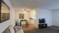This single-floor residence offers a warm and functional open-concept layout that brings together the living, dining, and kitchen areas in one cohesive space. The living room features plush carpeting and provides a comfortable setting for relaxation, anchored by a flat-screen TV setup on a sleek media console. A ceiling fan with an integrated light fixture overhead adds both practical airflow and ambient lighting to the combined living and dining zone. The dining area accommodates a compact bar-height table with stools, making it an ideal spot for casual meals. Decorative touches, including wall art and potted plants, add a homey character to the space. The kitchen is positioned just beyond the dining area and features white cabinetry, a white range, and light countertops, giving it a clean and bright appearance. A pass-through design between the kitchen and dining area enhances the sense of openness throughout. The textured ceiling and neutral wall tones create a consistent, understated palette that ties each area together. Overall, the layout is thoughtfully designed to maximize the use of space while maintaining a comfortable and inviting atmosphere throughout.