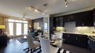 This single-floor residence showcases an open-concept layout that seamlessly connects the kitchen, dining, and living areas into one cohesive and functional space. The kitchen is outfitted with rich, dark cabinetry that reaches toward the ceiling, providing ample storage while contributing to the space's upscale aesthetic. Light-colored countertops create a striking contrast against the dark cabinets, and a classic subway tile backsplash adds texture and visual interest. Track lighting runs along the ceiling, illuminating the kitchen and dining areas with precision, while a ceiling fan with light fixture serves the living room.

The dining area features a glass-top table surrounded by elegant white upholstered chairs, anchored by a bold black-and-white geometric area rug. An accent wall painted in deep plum adds a dramatic focal point to the living area, which is furnished with a teal sofa and complementary décor. Large windows allow natural light to filter in, enhancing the warm and inviting ambiance. Dark hardwood-style flooring runs throughout, tying together the sophisticated, contemporary design of the entire space.