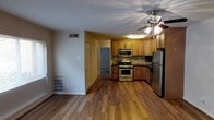 This single-floor residence offers a practical and well-designed open-concept layout that connects the living area and kitchen in a cohesive, functional space. The kitchen is a standout feature, outfitted with warm honey-toned wood cabinetry, a decorative tile backsplash, and a full suite of stainless steel appliances including a gas range, over-the-range microwave, and a full-size refrigerator. Ample upper and lower cabinetry provides generous storage throughout the kitchen. The open floor plan allows the kitchen to flow naturally into the main living area, which is anchored by rich, dark hardwood-style flooring that runs throughout the space, adding warmth and visual continuity. A ceiling fan with an integrated light fixture serves the main area, providing both air circulation and ambient lighting. The space also features visible HVAC vents, ensuring efficient climate control. Neutral wall tones keep the interior feeling bright and versatile. A doorway visible from the main living area leads to additional rooms, suggesting a well-connected interior layout that balances open communal spaces with private areas. Overall, the space offers a comfortable, functional environment with a cohesive design aesthetic.