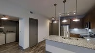 This single-floor space showcases a thoughtfully designed open-concept layout that seamlessly connects the kitchen and living areas. The kitchen serves as a focal point, anchored by a generous island topped with speckled granite countertops and an integrated sink, providing both a functional workspace and a casual gathering spot. A stainless steel refrigerator and classic subway tile backsplash complement the dark wood cabinetry, creating a balanced contrast between light and dark tones throughout the space.

Overhead, a combination of industrial-style pendant lights and track lighting suspended from the ceiling provides layered illumination, enhancing the modern aesthetic. The warm wood-look flooring runs continuously throughout, adding texture and cohesion to the open layout. Neutral white walls are paired with dark gray doors and trim, lending a contemporary, polished feel.

The entryway area is visible in the background, offering a clear sightline through the space, which contributes to an airy, open atmosphere. The overall design strikes a balance between modern sophistication and everyday practicality, with quality finishes and purposeful details evident throughout.