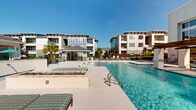 Located at 356 Lake Murray Boulevard in Columbia, South Carolina, this expansive outdoor amenity area serves as a centerpiece of a modern residential community. The space is defined by multiple swimming pools, including a large resort-style lap pool with crystal-clear blue water and a separate spa-style pool featuring decorative water jets along its edge. Surrounding the pools, a generously sized sundeck provides ample room for lounging, with in-pool tanning ledges outfitted with contemporary white chaise lounges and additional seating areas positioned throughout the patio. A wood-beamed pergola structure offers shaded outdoor seating alongside the main pool, complemented by cushioned sofas and accent furniture. A covered gazebo near the gated entrance provides additional shade and serves as a focal point for the secondary pool area. Lush tropical landscaping, including palm trees and colorful planters, enhances the resort ambiance throughout. The space is enclosed by ornamental iron fencing with a secured gate entry, ensuring privacy. The surrounding multi-story residential buildings, finished in coastal-inspired gray and blue siding with large windows and private balconies, frame the outdoor area and contribute to the cohesive, upscale aesthetic of the community.