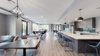 This contemporary single-floor community lounge is thoughtfully designed to offer a versatile and welcoming environment for gathering, socializing, and relaxing. The open-plan layout is anchored by a striking, oversized kitchen island with a crisp white countertop and dark cabinetry, surrounded by sleek navy blue bar stools. Pendant lighting in brushed gold and matte black tones hangs above the island, adding a refined touch to the space. The fully equipped kitchen area features stainless steel appliances, a tiled backsplash in deep blue-green, ample upper cabinetry, and a wall-mounted television. Light wood-tone flooring flows seamlessly throughout, complementing the cool grey walls and white trim. Multiple high-top bar tables with modern black chairs are positioned near large windows and glass doors, inviting an abundance of natural light and offering pleasant views of the surrounding greenery. A billiards table is tucked toward the back of the room, providing a recreational focal point. A separate lounge nook offers comfortable seating for more intimate conversation. Recessed lighting and statement pendants ensure the space is well-lit throughout, creating a bright, modern, and inviting atmosphere ideal for both casual gatherings and communal activities.