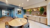 Located at 18750 Delaware St in Huntington Beach, California, this thoughtfully designed community space offers a welcoming and functional environment across a single floor. The dining area serves as the centerpiece, featuring a round natural wood dining table surrounded by upholstered chairs, all bathed in recessed lighting and positioned beside expansive floor-to-ceiling sliding glass doors that open directly to a landscaped outdoor courtyard — flooding the interior with natural light and seamless indoor-outdoor connectivity.

The kitchen runs along one wall with light wood cabinetry, a white quartz countertop, a gold-accented sink, a built-in oven, and open shelving adorned with lush greenery and decorative objects. A textured mosaic tile backsplash adds warmth and visual interest. Bold blue accent walls and curated artwork, including a vibrant botanical print, lend the space a contemporary, artistic character.

A cozy lounge nook features a mustard velvet sofa, terracotta accent chairs, and layered wall sconces set against patterned wallpaper. Light terrazzo-style flooring ties the dining and lounge zones together cohesively.

A carpeted corridor connects the space to private glass-enclosed rooms — ideal for meetings or focused work — and a coworking suite, making this a versatile, amenity-rich gathering destination.