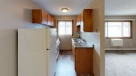 This single-floor unit offers a practical and functional layout designed for comfortable everyday living. The galley-style kitchen is lined with warm, medium-toned wood cabinetry on both upper and lower levels, providing ample storage space. Dark granite-style countertops complement the cabinetry, while a double-basin sink sits beneath a window fitted with vertical blinds, allowing natural light to filter in. White appliances, including a refrigerator and range, complete the kitchen's utilitarian setup. The flooring transitions from hardwood-style planks in the kitchen and entryway to plush carpet throughout the main living areas. The spacious living room is characterized by neutral taupe walls, a textured ceiling, and a large picture window that brings in additional natural light. A baseboard heating unit runs along the exterior wall beneath the window. The entry foyer features a solid wood door and a clear transition point between the hard flooring and carpeted areas. A hallway connects the living spaces to the bathroom, which is visible at the corridor's end, keeping the layout straightforward and easy to navigate. Overall, the space presents a clean, understated aesthetic with warm wood accents.