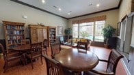 This single-floor library is thoughtfully designed to serve as both a reading retreat and a social gathering space. The room features warm hardwood flooring throughout, complemented by soft sage-green walls accented with dark wainscoting that adds a refined, traditional character. Crown molding and recessed lighting contribute to the polished architectural finish overhead.

Bookshelves—both freestanding pine armoires and open bookcases stocked with a wide collection of titles—line the walls, providing ample reading material. The space is divided into distinct functional areas: a reading and lounge zone anchored by a tan upholstered sofa and accent chairs arranged around a glass-topped coffee table, and a separate seating area with ornate dark wood round tables and high-back chairs, suitable for group activities or casual dining.

Large windows dressed with decorative fabric valances flood the space with natural light and frame outdoor views of lush greenery, palm trees, and surrounding architecture. Framed artwork and potted indoor trees add warmth and visual interest. An adjacent room with additional dining seating and a service area is visible through a wide opening, extending the overall functionality of the space.