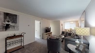 This single-floor residence offers a thoughtfully designed layout that balances comfort and functionality across its key living areas. The living room serves as the heart of the home, featuring plush carpeting, neutral warm-toned walls, and generous space for seating arrangements. A built-in closet area and open sightlines contribute to the room's airy feel. Adjacent to the living space, a cozy dining nook is highlighted by a modern chandelier, textured accent wall, and a window that invites natural light, creating an intimate setting for meals. The kitchen is efficiently arranged in a U-shaped configuration, showcasing rich dark cabinetry, granite-style countertops, wood-look flooring, and white appliances including a range, refrigerator, and microwave. The bedroom is a serene retreat, characterized by soft carpeting, warm gray walls, and large windows that frame outdoor greenery, flooding the space with natural light. A ceiling fixture and ample room for furnishings round out this restful space. The bathroom features a generously sized vanity with dark wood cabinetry, a light countertop, and a large mirror, providing practical storage and a clean, polished finish throughout.