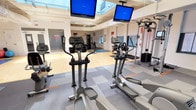 This fitness center is a spacious, single-floor facility designed to accommodate a wide range of workout activities. The space is defined by its notably high ceilings, which are accentuated by a large central skylight that floods the open floor area with natural light, creating a bright and energizing environment. University banners hang from the upper walls, adding an institutional character to the space.

The cardio zone is well-stocked with elliptical trainers, treadmills, stationary bikes, and a rowing machine, all arranged on interlocking rubber-tiled flooring with orange accent strips. Multiple flat-screen monitors are mounted from the ceiling above the cardio equipment, providing entertainment during workouts. A dedicated strength training area offers a cable and pulley multi-station machine, free weights, and additional resistance equipment positioned along the perimeter.

A large open section of hardwood flooring serves as a functional training and stretching area, equipped with exercise mats and stability balls. Full-length mirrors line one wall, expanding the sense of space. The facility also includes a separate enclosed room, clearly marked as a Fitness Center entrance, along with additional workout areas visible through glass partitions, providing distinct zones for varied training needs.