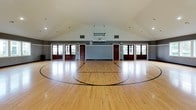 This impressive indoor sports facility centers around a full-sized basketball court with a beautifully finished hardwood floor, complete with clearly marked court lines for basketball and additional multi-sport use. The space is defined by its dramatic vaulted ceiling, which rises to a generous peak and is fitted with recessed lighting that evenly illuminates the entire court. A regulation basketball hoop is mounted and suspended from the ceiling structure, ready for play.

The walls feature a two-tone design, with a light beaded wainscoting panel along the lower half and a bold dark gray upper section, giving the space a polished, finished look. Multiple large windows are positioned along the walls, inviting an abundance of natural light and offering pleasant views of the surrounding greenery outdoors.

Access to the court is provided through a set of stately, dark wood-framed French-style doors with glass pane inserts, lending a refined architectural character to what is otherwise a purely athletic environment. Additional interior doors provide access to adjacent rooms or utility spaces. The overall ambiance is clean, open, and well-appointed, making this facility equally suited for competitive play, recreation, and fitness activities.