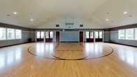 This impressive indoor sports facility centers around a full-sized basketball court with a beautifully finished hardwood floor, complete with clearly marked court lines for basketball and additional multi-sport use. The space is defined by its dramatic vaulted ceiling, which rises to a generous peak and is fitted with recessed lighting that evenly illuminates the entire court. A regulation basketball hoop is mounted and suspended from the ceiling structure, ready for play.

The walls feature a two-tone design, with a light beaded wainscoting panel along the lower half and a bold dark gray upper section, giving the space a polished, finished look. Multiple large windows are positioned along the walls, inviting an abundance of natural light and offering pleasant views of the surrounding greenery outdoors.

Access to the court is provided through a set of stately, dark wood-framed French-style doors with glass pane inserts, lending a refined architectural character to what is otherwise a purely athletic environment. Additional interior doors provide access to adjacent rooms or utility spaces. The overall ambiance is clean, open, and well-appointed, making this facility equally suited for competitive play, recreation, and fitness activities.