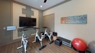 The Aerobics Center is a thoughtfully designed fitness space finished with warm wood-look flooring and neutral-toned walls that create an inviting, energetic atmosphere. The room is anchored by a large flat-screen television mounted on a decoratively paneled accent wall with subtle geometric detailing, making it ideal for guided workout sessions and cycling classes. Several spin bikes are arranged facing the screen, providing an immersive cycling experience for group or solo training.

Natural light filters into the space through a large window fitted with horizontal blinds, offering outdoor views while maintaining a bright, open feel. A ceiling fan overhead provides comfortable air circulation during intense workouts. The room is equipped with an impressive array of fitness accessories, including a tiered dumbbell rack, a full set of SPRI weighted bars on a dedicated stand, medicine balls displayed on a vertical rack, resistance bands, stability balls, step platforms, and weighted bags — offering ample options for full-body training. A glass-paneled door provides easy access between areas of the facility. The combination of well-organized equipment, decorative touches, and practical amenities makes this aerobics center a functional and welcoming space for diverse fitness routines.