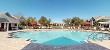 This inviting outdoor pool area is thoughtfully designed to offer a resort-like experience. At its centerpiece is a generously sized swimming pool with crystal-clear blue water, framed by a well-appointed paver deck that provides plenty of room for lounging. Sun chairs and reclining loungers are arranged throughout the deck, ideal for soaking up the sunshine, while a shaded pergola and large red market umbrellas offer welcome relief from the sun.

Adjacent to the pool, a covered patio features classic white columns, a beadboard ceiling, and a brick-accented exterior wall with arched transom windows. This sheltered area is furnished with bold red cushioned sofas and a metal coffee table, creating a comfortable outdoor living space perfect for relaxing and socializing.

Decorative potted plants add greenery and warmth throughout the patio, while the surrounding landscape includes mature trees that provide a natural backdrop. The pool deck's stone pavers extend seamlessly between the covered lounge and the water's edge, tying the entire space together with a cohesive, polished aesthetic.

Beyond the pool, scenic waterfront views and lush landscaping complete the setting, making this outdoor amenity a standout feature of the community.
