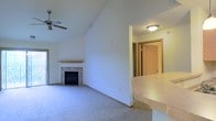 This single-floor residence offers a thoughtfully designed layout that balances open living with functional flow. The living room serves as the heart of the home, drawing the eye upward with its vaulted ceiling that lends a sense of spaciousness and airiness to the space. A ceiling fan is centrally mounted above, providing both comfort and a subtle design detail. The room's focal point is a tile-surround fireplace, complete with a light wood mantle, adding warmth and character to the area. Soft, plush carpeting throughout the living space contributes to a cozy and comfortable atmosphere. Adjacent to the living room, a raised counter or breakfast bar provides a natural transition into the kitchen area, offering both a casual dining or workspace surface. Light-toned laminate countertops complement the neutral color palette maintained throughout the home. A hallway visible from the main living area leads to additional rooms, with natural wood-toned doors adding warmth to the interior. The overall design makes use of clean white walls and consistent neutral tones, creating a bright and welcoming environment that is both versatile and easy to personalize.