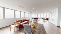 Located at 104 W Franklin St in Richmond, VA, this generously sized single-floor community living space offers an open, airy atmosphere defined by wraparound floor-to-ceiling windows that flood the interior with natural light and frame sweeping views of the Richmond skyline and a rooftop pool area beyond. The light wood-tone flooring runs throughout, complementing the clean white walls and creating a bright, modern aesthetic.

The space is thoughtfully divided into multiple functional zones. A primary lounge area features a rich cognac leather sofa paired with contemporary gray accent chairs, all anchored by a patterned area rug. A secondary seating zone offers more casual furniture arranged to take advantage of the panoramic window views. A dedicated recreation area houses a full-size pool table and bar-height seating, perfect for social gatherings.

Along one wall, built-in media storage units support a wall-mounted flat-screen television, while classic crown molding adds an elegant architectural touch. Recessed and linear ceiling lighting supplements the ample daylight. The open-plan layout, supported by a central structural column, allows for flexible use of the space while maintaining a cohesive, inviting ambiance throughout.