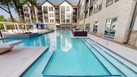 The pool area is a expansive, resort-inspired outdoor amenity space situated within a modern multi-story residential community. The centerpiece is a large, rectangular swimming pool with clear turquoise water, clean geometric lines, and a raised integrated spa or wading feature. Surrounding the pool is a generous paved deck finished in light-toned concrete, providing ample room for lounging and entertaining. Multiple padded chaise lounge chairs are positioned along the pool's perimeter, offering residents convenient sunbathing areas. A standout feature of the space is the presence of bold blue cabanas, each equipped with flowing drape curtains that can be drawn for shade and privacy. Inside these cabanas, additional chaise lounges and accent tables create comfortable, semi-private retreats. A white pergola structure provides an additional shaded gathering space on the far end of the deck. Planter beds with greenery are integrated into the hardscape, adding natural texture to the environment. The residential building's stone and panel facade frames the pool on multiple sides, while a natural tree line borders the outer perimeter, enhancing privacy and providing a lush, green backdrop to the overall space.