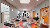 Located at 14437 SW Teal Blvd in Beaverton, Oregon, this single-floor fitness center combines modern design with a comprehensive range of workout equipment. The space is immediately distinguished by its eye-catching chevron-patterned flooring in vivid shades of orange, red, and gray, adding a dynamic energy to the environment. Overhead, large skylights flood the interior with natural light, complemented by recessed ceiling fixtures and wall-mounted sconces that ensure consistent illumination throughout.

The cardio zone is well-appointed with multiple treadmills, elliptical machines, and stationary bikes from brands including True and Cybex, all arranged to maximize usable floor space. A wall-mounted flat-screen television provides entertainment during workouts. The strength training area features a dedicated free weights section with a full dumbbell rack, resistance machines, and a Cybex strength system, along with exercise balls and additional functional fitness equipment.

Large windows and a glass-paneled entry door with a bold orange frame offer views of the surrounding outdoor landscape, reinforcing a sense of openness. Bold orange doors lead to additional rooms, and a leather lounge chair provides a rest area. Full-length mirrors along one wall enhance the sense of space and support proper form during training sessions.