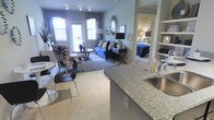 This single-floor residence is designed around an open-concept layout that seamlessly connects the kitchen, dining, and living areas, creating a cohesive and functional living space. The kitchen anchors the foreground with a granite-style countertop peninsula and a stainless steel sink, offering both workspace and a natural visual divider between the cooking and living zones. Open shelving nearby provides additional display and storage options.

The dining area accommodates a compact round table paired with stylish black chairs, making efficient use of space without feeling cramped. Beyond it, the living room is bright and inviting, benefiting from multiple windows dressed with bold navy and white patterned curtains and valances that add a strong design accent. A vibrant blue sofa, decorative throw pillows, and a patterned area rug contribute to the room's lively aesthetic, while a sunburst mirror on the wall adds an eye-catching focal point.

A hallway leads to a private bedroom, visible in the background, which features warm lamp lighting and a blue-accented bedspread, maintaining the cohesive color palette found throughout the space. Neutral toned flooring and light gray walls provide a balanced, modern backdrop throughout.