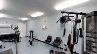 This single-floor fitness space offers a clean, functional environment designed for exercise and physical activity. The room features light grey walls and a smooth grey floor, creating a neutral, no-frills atmosphere that keeps the focus on fitness. Overhead fluorescent panel lighting ensures the space is well-illuminated throughout.

The room is equipped with a multi-station cable gym machine prominently positioned to one side, featuring pull-down bars and adjustable components suitable for a variety of strength training exercises. A stationary exercise bike occupies the opposite corner, offering a dedicated option for cardiovascular workouts. A small wall-mounted shelf provides a convenient spot for personal items or reading material during exercise sessions.

Two black chairs are placed along the wall, offering a rest area between sets or a place for observers. A mounted screen above the multi-station unit adds an entertainment or instructional element to the workout experience. The space is accessed through a standard white door, and the overall layout is efficient and straightforward, making it a practical fitness facility for daily use.