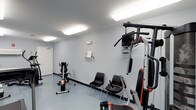 This single-floor fitness space offers a clean, functional environment designed for exercise and physical activity. The room features light grey walls and a smooth grey floor, creating a neutral, no-frills atmosphere that keeps the focus on fitness. Overhead fluorescent panel lighting ensures the space is well-illuminated throughout.

The room is equipped with a multi-station cable gym machine prominently positioned to one side, featuring pull-down bars and adjustable components suitable for a variety of strength training exercises. A stationary exercise bike occupies the opposite corner, offering a dedicated option for cardiovascular workouts. A small wall-mounted shelf provides a convenient spot for personal items or reading material during exercise sessions.

Two black chairs are placed along the wall, offering a rest area between sets or a place for observers. A mounted screen above the multi-station unit adds an entertainment or instructional element to the workout experience. The space is accessed through a standard white door, and the overall layout is efficient and straightforward, making it a practical fitness facility for daily use.