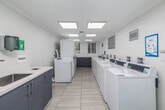 Located at 8665 Burton Way in Los Angeles, CA, this shared laundry facility offers a clean, well-organized environment designed for efficient and convenient use. The space features light-toned tile flooring throughout, crisp white walls, and crown molding details that lend a polished appearance. Bright flush-mount panel lighting and recessed ceiling lights ensure the room is well-lit at all hours.

The laundry area is equipped with multiple top-load washing machines and stacked dryer units, all finished in white and arranged in parallel rows to maximize capacity. Each machine is fitted with a digital control panel, and dedicated utility hookup stations are positioned along the walls. A utility sink with gray cabinetry and a white countertop provides a practical space for hand-washing or pre-treatment, complemented by mounted soap and paper towel dispensers.

An adjacent anteroom connects to the facility and features striking decorative wall graphics with Beverly Hills-themed imagery, alongside smart package locker units — adding both visual interest and functional amenity to the space. Posted signage clearly outlines laundry room rules and operating instructions. Windows at the end of the laundry room allow natural light to filter in, offering views of surrounding greenery and enhancing the overall brightness and comfort of the facility.