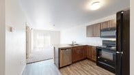 Located at 1330 Catron Blvd in Rapid City, South Dakota, this single-floor unit offers a practical and comfortable layout designed for everyday living. The open-concept kitchen and living area form the heart of the space, with the kitchen featuring rich, warm-toned cabinetry, a peninsula with a double sink and light countertops, and black appliances including a range, over-the-range microwave, refrigerator, and a stainless steel dishwasher. Wood-look flooring runs throughout the kitchen and connects seamlessly to the adjoining living area, which is finished with plush carpet and receives natural light through a sliding vertical-blind window treatment. The bedroom is a calm, carpeted retreat with a window offering natural light and a modern wood-grain door. The bathroom is finished with wood-look flooring, a full tub and shower combination, a toilet, and a vanity. A dedicated laundry room provides added convenience with a full-size washer and dryer and built-in shelving. Throughout the unit, neutral walls, flush-mount ceiling lighting, and clean white trim contribute to a bright, move-in-ready atmosphere well-suited for comfortable residential living.