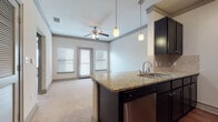 This single-floor, one-bedroom, one-bathroom unit is thoughtfully designed to maximize functionality within a compact footprint. The open-concept layout seamlessly connects the kitchen and living area, creating a cohesive and practical living space. The kitchen is equipped with dark espresso cabinetry, granite countertops, a tile backsplash, and stainless steel appliances including a refrigerator, microwave, and dishwasher. Pendant lighting over the kitchen peninsula adds a stylish focal point while providing task lighting. The living area features plush carpet flooring, a ceiling fan, and access to a private balcony through glass-panel doors flanked by windows that bring in natural light. The bedroom is carpeted and provides a quiet, versatile space. The bathroom offers a granite-topped vanity with dark cabinetry, a soaking tub with subway tile surround, and wood-look flooring for a polished finish. A spacious walk-in closet with wire shelving and washer/dryer connections adds practical storage and convenience throughout the unit.