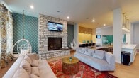 Located at 7220 E Broad St in Columbus, Ohio, this single-story community clubhouse offers a thoughtfully designed amenity space with distinct functional zones. The main lounge serves as the centerpiece, featuring a striking floor-to-ceiling natural stone fireplace with a mounted flat-screen TV, teal diamond-quilted accent wall, plush sofas, and a vibrant Persian-style rug anchoring the seating area. Natural light filters in through large windows dressed with floral and geometric drapery, adding warmth to the space. Adjacent to the lounge, a well-appointed kitchen area features white shaker cabinetry, quartz countertops, a dark tile backsplash, stainless steel appliances, and a counter-height bar with woven wishbone stools beneath brass pendant lighting. A business center with computer workstations sits under warm pendant lights against a yellow accent wall, and a billiards table occupies a separate recreational zone. A cozy reading nook with deep navy walls, ambient sconce lighting, and a large window provides a quiet retreat. The herringbone-patterned light wood flooring flows throughout, and glass-paneled sliding barn doors and glass partition walls contribute to an open, modern aesthetic with views toward an outdoor pool area.