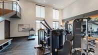Located in Fort Worth, TX, this two-floor fitness center offers a generously sized, well-appointed exercise facility designed to support a wide range of workout routines. The ground floor makes an immediate impression with its dramatic double-height ceilings and floor-to-ceiling windows that flood the space with natural light while offering outdoor views. This level is outfitted with a comprehensive array of True-brand cardio equipment, including treadmills and ellipticals arranged in a row along the windows, as well as a full selection of cable and strength-training machines positioned on durable, patterned carpet flooring. A built-in entertainment console with a TV and organized weight storage adds functional detail to the lower level. A sleek steel staircase with warm wood accents connects to the upper floor, which transitions to light hardwood flooring and features a dedicated studio-style exercise room. This upper level is equipped with stationary bikes, rowing machines, wall-to-wall mirrors, stability balls, foam rollers, and free weights, making it ideal for cycling sessions, yoga, or group fitness. Recessed lighting and ceiling fans maintain a comfortable, energizing atmosphere throughout. The modern design, clean white walls, and thoughtful layout across both levels create a professional and motivating environment suited for a comprehensive fitness experience.