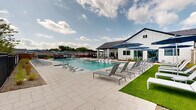 Located at 151 S Buddy Hayes Blvd in Anna, Texas, this single-level community amenity facility offers an impressively spacious outdoor experience centered around a large, resort-style swimming pool. The pool area is finished with broad, light-toned stone pavers that extend generously around all sides, providing ample room for lounging and socializing. Rows of modern sling-style chaise lounges line the sun deck, accompanied by large cantilever umbrellas in a classic navy blue that offer shaded retreats throughout the day.

The adjacent clubhouse building is clad in crisp white board-and-batten siding accented by a dark pitched roof, reflecting a contemporary farmhouse aesthetic. A covered outdoor pavilion extends from the building, equipped with ceiling fans and furnished with dining tables and chairs, creating an ideal space for gatherings in any weather. The pool itself features in-water tanning ledges with additional loungers, perfect for relaxing at the water's edge.

Decorative brick columns punctuate the perimeter, complemented by manicured landscaping including ornamental grasses and young trees that add greenery and texture to the hardscape. A separate pergola structure on the far end of the deck provides an additional shaded seating area. The entire space is enclosed by black iron fencing, offering both security and an open, airy feel under the expansive Texas sky.