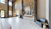 Located at 202 Park Blvd in Cherry Hill, NJ, this sophisticated lobby space makes an immediate impression with its dramatic double-height ceilings and expansive, open floor plan. Large-format marble-look porcelain tiles stretch across the floor, reflecting abundant natural light that pours in through tall windows and glass entry doors. A bold, sculptural sputnik-style chandelier with gold and matte black accents anchors the central ceiling, adding a contemporary artistic focal point to the space.

At the heart of the lobby sits a generously sized, curved tufted seating piece upholstered in white, wrapped in a warm caramel leather base — an elegant and inviting centerpiece. A full-height mirrored wall panel with gold-trimmed dark columns amplifies the sense of depth and grandeur, visually expanding the already spacious interior.

The entry vestibule features floor-to-ceiling glazed doors framed in black, allowing clear views to the exterior and filling the space with daylight. Adjacent areas include a mail room with secure mailboxes and a furnished lounge area with blue accent chairs, providing both functional and comfortable spaces for occupants. The overall ambiance is modern and polished, blending warm neutral tones with metallic accents to create a welcoming yet refined environment.