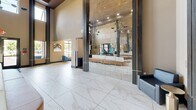 Located at 202 Park Blvd in Cherry Hill, NJ, this sophisticated lobby space makes an immediate impression with its dramatic double-height ceilings and expansive, open floor plan. Large-format marble-look porcelain tiles stretch across the floor, reflecting abundant natural light that pours in through tall windows and glass entry doors. A bold, sculptural sputnik-style chandelier with gold and matte black accents anchors the central ceiling, adding a contemporary artistic focal point to the space.

At the heart of the lobby sits a generously sized, curved tufted seating piece upholstered in white, wrapped in a warm caramel leather base — an elegant and inviting centerpiece. A full-height mirrored wall panel with gold-trimmed dark columns amplifies the sense of depth and grandeur, visually expanding the already spacious interior.

The entry vestibule features floor-to-ceiling glazed doors framed in black, allowing clear views to the exterior and filling the space with daylight. Adjacent areas include a mail room with secure mailboxes and a furnished lounge area with blue accent chairs, providing both functional and comfortable spaces for occupants. The overall ambiance is modern and polished, blending warm neutral tones with metallic accents to create a welcoming yet refined environment.