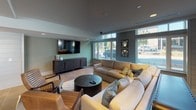 Located at 2–12 Perry Ave in Greenville, SC, this single-floor community amenity space offers a well-appointed, multi-functional environment designed for social gathering and leisure. The layout encompasses several distinct zones that flow seamlessly into one another, all finished with large-format tile flooring and a cohesive neutral color palette of grays, whites, and warm tones.

The entrance foyer welcomes visitors with floor-to-ceiling glass double doors that invite abundant natural light and offer views of the surrounding streetscape. Two upholstered armchairs and nature-inspired wall art create an inviting first impression. Adjacent to the entry, a lounge area features tufted leather seating arranged around an oversized Scrabble board display and chalkboard wall, adding a playful, interactive element to the space.

A dedicated game room includes a round poker table with seating for several players, accompanied by a wall-mounted television and a window overlooking an outdoor patio. Nearby, a spacious TV lounge offers plush sectional and accent seating oriented toward a large flat-screen, with expansive windows flooding the room with natural light.

The dining and social area is anchored by a substantial dark wood communal table beneath striking globe pendant fixtures, complemented by built-in cabinetry, a mini-refrigerator, and a mounted television — ideal for casual gatherings or events. Together, these areas form a versatile, modern community hub.