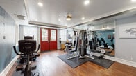 This single-level fitness center offers a thoughtfully designed workout space that balances functionality with a polished aesthetic. The room features warm hardwood flooring throughout, accented by protective rubber mats beneath the equipment stations. A tray ceiling with crown molding adds architectural character, while recessed lighting and a ceiling fan ensure the space remains well-lit and comfortable during workouts.

A bold set of red double doors serves as the main entrance, flanked by windows dressed with white blinds that allow natural light to filter in and provide views of the surrounding outdoor area. An entire wall of floor-to-ceiling mirrors visually expands the space and allows users to monitor their form during exercise.

The gym is comprehensively equipped with a multi-station cable weight machine, a treadmill, an elliptical, an upright stationary bike, a recumbent bike, and a flat bench. A wall-mounted television is positioned for easy viewing from multiple exercise stations. Additional accessories, including kettlebells, resistance bands, and rolled towels, are neatly organized on a storage shelf. Motivational signage and artwork decorate the walls, contributing to an energizing and focused training environment.