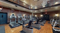 This expansive fitness facility combines industrial architectural character with a comprehensive range of workout equipment. The space is defined by its dramatic exposed brick walls, soaring double-height ceilings, and a wraparound mezzanine level enclosed by metal railings — giving the gym a grand, loft-like atmosphere. Natural light filters in through large multi-pane windows along the upper level, complemented by warm pendant and track lighting that creates an energizing yet welcoming ambiance throughout.

The main floor features classic hardwood flooring across a generous open area, accommodating multiple distinct workout zones. A robust cardio section houses a full row of treadmills equipped with individual display screens, alongside recumbent and upright stationary bikes and a dedicated spin cycling area. The strength training zone is well-appointed with a variety of cable machines, weight benches, squat racks, and a full dumbbell rack. An open floor area provides ample room for functional training, stretching, and group fitness activities, complete with mats and freestanding equipment. Exposed ductwork, black steel columns, and industrial-style doors add to the raw, urban aesthetic, making this a visually distinctive and fully equipped fitness destination.