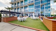 Located at 215 N Pine Street in the heart of downtown Charlotte, NC, this outdoor amenity deck offers a well-appointed rooftop retreat with an impressive array of features designed for both relaxation and social gathering. The space is defined by a generous open-air layout finished with large-format stone-look pavers that lend a clean, modern aesthetic throughout.

A lush turf lawn area provides a welcoming green focal point, surrounded by warm wood-trimmed raised planters filled with colorful flowers and tropical palm trees. White lattice-back chairs and round tables are arranged across the turf, creating an inviting setting for casual dining and outdoor socializing.

A sleek steel pergola structure anchors the grilling zone, which features multiple stainless steel grills set against stone-clad outdoor kitchen stations — ideal for resident gatherings and events. String lights are strung beneath the pergola, adding ambiance for evening use.

The deck is fully enclosed by perimeter metal fencing, ensuring safety while preserving unobstructed panoramic views of the Charlotte skyline, with high-rise towers visible in multiple directions. Tall light poles provide ample illumination throughout the space after dark. The overall design balances urban sophistication with functional outdoor living, making it a standout amenity in the downtown Charlotte residential landscape.