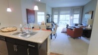 This single-floor residence features an open-concept layout that seamlessly integrates the kitchen, dining, and living areas into one cohesive space. The kitchen is appointed with granite countertops, dark espresso cabinetry, a double-basin stainless steel sink, and a peninsula island that provides additional prep space and casual seating. A pendant light fixture hangs above the island, adding a stylish focal point. The adjacent dining area accommodates a full dining table with seating for multiple guests, complemented by decorative artwork on the wall. The living area extends naturally from the dining space, featuring plush carpeting, a seating arrangement with accent chairs, and a patterned area rug that defines the lounge zone. Large windows throughout the space allow abundant natural light to pour in, creating a bright and airy ambiance. Sheer curtains and roman shades offer both privacy and light control. The overall design is modern and inviting, with a neutral color palette accented by warm tones, contributing to a welcoming and functional living environment.