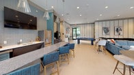 Located at 6925 Roswell Rd in Sandy Springs, GA, this single-floor clubhouse is a thoughtfully designed amenity space that combines functionality with contemporary style. The open-plan layout is anchored by a well-appointed kitchen area featuring stainless steel appliances, a striking granite-topped island with seating for several guests, and teal-accented tile backsplash that adds a bold visual contrast to the neutral palette. Elegant pendant lighting with glass fixtures hangs overhead, complementing the recessed lighting throughout the space. Large-format tile flooring in a soft, warm tone runs seamlessly across the entire floor, reinforcing the spacious feel. A dedicated lounge area offers generous sectional seating in cool blue upholstery, centered around a large wall-mounted display screen — ideal for group gatherings or media viewing. A billiards table provides additional entertainment options toward the rear of the space. Floor-to-ceiling windows dressed with sheer linen curtains flood the interior with natural light while offering direct views of the outdoor pool area and surrounding landscaped grounds. Curated modern artwork and layered seating arrangements — from bar-height tables to intimate conversation nooks — ensure the space is versatile and inviting for a range of social occasions.