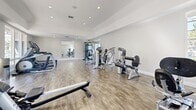 Located at 655 Baker St in Costa Mesa, California, this fitness center offers a well-equipped exercise space designed to support a comprehensive workout routine. The room features wood-style flooring throughout, crisp white walls, and a tray ceiling with recessed lighting that creates a bright, clean atmosphere. Large windows and glass doors line the perimeter, flooding the space with abundant natural light and providing views of the surrounding greenery and outdoor areas. The cardio zone includes multiple treadmills, an elliptical trainer, and a Matrix rowing machine, all positioned to take advantage of the natural light and outdoor views. A dedicated strength training area houses several selectorized resistance machines, including a lat pulldown, chest press, and fly/rear delt machine, offering a solid range of upper and lower body exercise options. A large wall-mounted mirror expands the sense of space and allows users to monitor their form during workouts. The overall ambiance is clean, airy, and modern, making for an inviting environment conducive to regular fitness activity. The space is thoughtfully arranged to maximize functionality while maintaining an open, uncluttered feel throughout.
