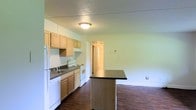 This single-floor residence offers a practical and functional layout designed for comfortable everyday living. The kitchen serves as a central feature of the home, equipped with light wood cabinetry that provides ample storage space. White appliances, including a refrigerator, electric range, dishwasher, and a double-basin sink, are neatly arranged along the countertop wall, complemented by dark laminate countertops that contrast cleanly with the light cabinetry. A freestanding kitchen island with a dark countertop surface adds additional workspace and serves as a natural divider between the kitchen and the adjoining living area. Rich, dark hardwood flooring runs throughout, anchoring the space with warmth and visual depth. The walls are finished in a soft, neutral tone that enhances the brightness of the interior, while a textured ceiling adds a subtle layer of character. A flush-mount ceiling light provides overhead illumination in the kitchen. A hallway visible from the kitchen leads to additional rooms, suggesting a well-connected and easy-to-navigate floor plan. Overall, the space presents a clean, no-frills aesthetic that balances functionality with a welcoming atmosphere.