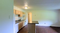 This single-floor residence offers a practical and functional layout designed for comfortable everyday living. The kitchen serves as a central feature of the home, equipped with light wood cabinetry that provides ample storage space. White appliances, including a refrigerator, electric range, dishwasher, and a double-basin sink, are neatly arranged along the countertop wall, complemented by dark laminate countertops that contrast cleanly with the light cabinetry. A freestanding kitchen island with a dark countertop surface adds additional workspace and serves as a natural divider between the kitchen and the adjoining living area. Rich, dark hardwood flooring runs throughout, anchoring the space with warmth and visual depth. The walls are finished in a soft, neutral tone that enhances the brightness of the interior, while a textured ceiling adds a subtle layer of character. A flush-mount ceiling light provides overhead illumination in the kitchen. A hallway visible from the kitchen leads to additional rooms, suggesting a well-connected and easy-to-navigate floor plan. Overall, the space presents a clean, no-frills aesthetic that balances functionality with a welcoming atmosphere.