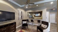 This thoughtfully designed single-floor space exudes a warm, sophisticated ambiance through its cohesive blend of neutral tones, refined furnishings, and architectural detail. The main living area features a tray ceiling accented with crisp white crown molding, a centrally mounted ceiling fan, and recessed lighting that creates a bright and inviting atmosphere. Rich taupe walls provide a warm backdrop for dark-framed artwork and contemporary décor, while tile flooring grounds the space with a natural, earthy texture. A patterned area rug anchors the seating arrangement, which includes dark upholstered accent chairs with contrasting decorative pillows, adding a touch of elegance. A wall-mounted flat-screen television and a classic dark wood console complete the lounge area. The open wet bar is a standout feature, boasting a granite countertop with bar seating, pendant lighting, a large framed mirror, a small refrigerator, and a sink — all framed within a pass-through opening that seamlessly connects the spaces. Decorative floral arrangements and carefully chosen accessories add a polished, finished look throughout. The overall design balances comfort and style, making this a welcoming and functional space ideal for relaxing or entertaining.