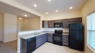 This single-story residence at Windhurst Park offers a thoughtfully designed layout with a clean, modern aesthetic throughout. The kitchen serves as a functional centerpiece, featuring dark espresso-toned cabinetry with ample upper and lower storage, granite-style countertops, and a full suite of black appliances including a refrigerator, range, built-in microwave, and dishwasher. Light wood-look flooring runs through the kitchen, which also benefits from recessed lighting and a door providing direct outdoor access alongside a window that draws in natural light. A breakfast bar-style counter connects the kitchen to the living area, creating an open, communicative flow between spaces.

The living area is generously sized, finished with plush carpet and a ceiling fan overhead. Bedrooms continue the carpeted flooring and ceiling fan combination, with windows offering outdoor views. A dedicated laundry room includes washer and dryer hookups, wire shelving, and tile flooring for practicality. The bathroom features a tub-shower combination, a granite-style vanity countertop with an undermount sink, a large mirror, and matching tile flooring, completing a cohesive and functional interior.
