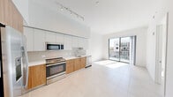 Located at 999 NW Seventh St in Miami, FL, this one-bedroom, one-bathroom unit offers a clean, contemporary design throughout its efficiently organized single-floor layout. The open-concept living and kitchen area is immediately striking, featuring soaring ceilings and large-format light-toned porcelain tile flooring that extends seamlessly across the space. The kitchen is outfitted with a full suite of stainless steel appliances — including a French-door refrigerator, electric range, built-in microwave, and dishwasher — complemented by warm wood-toned lower cabinetry, crisp white upper cabinets, a subway tile backsplash, and a white quartz countertop. Track lighting overhead adds a modern, functional touch. The living area is bathed in natural light through a floor-to-ceiling sliding glass door that opens to an outdoor-facing view, enhancing the airy ambiance of the space. The bedroom is comfortably sized with large windows overlooking the surrounding Miami cityscape, and features a sliding barn-style closet door. The bathroom is finished with matching wood-toned cabinetry, an undermount sink, and a glass-enclosed walk-in shower, maintaining the cohesive modern aesthetic found throughout the unit. A dedicated closet and entrance area complete the thoughtfully designed floor plan.