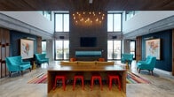 This dynamic communal lounge is designed with an industrial-modern aesthetic that blends raw materials with bold, colorful accents. The centerpiece of the main gathering area is a striking dark brick feature wall, anchored by a linear gas fireplace and a wall-mounted flat-screen TV, with a dramatic cluster of Edison bulb pendants suspended from the soaring double-height ceiling above. Floor-to-ceiling windows on multiple sides flood the space with natural light and offer expansive views of the surrounding cityscape, while sliding glass doors provide seamless access to an outdoor terrace. Polished concrete floors run throughout, complemented by vivid, multi-striped area rugs that add warmth and visual energy. Seating is thoughtfully varied, including a long communal wood table with bold red metal stools, lounge chairs in teal and chartreuse upholstery, and intimate conversation groupings separated by slatted wood screens. A wet bar area features a vibrant mosaic tile backsplash and is illuminated by oversized black pendant lights, making it ideal for social gatherings. A billiards table occupies a dedicated game zone, further enhancing the recreational appeal. Exposed ductwork and concrete structural elements underscore the loft-inspired character of the space, resulting in an environment that is both energetic and inviting.