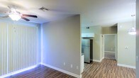 This single-floor space offers a clean, open layout designed for comfortable everyday living. The living area is bathed in natural light streaming through vertical-blind-dressed windows, creating a bright and welcoming atmosphere. Warm wood-style plank flooring runs throughout, adding a sense of continuity and warmth to the space.

The kitchen is efficiently arranged and features stainless steel appliances, including a full-size refrigerator, paired with crisp white upper cabinetry that provides ample storage. The kitchen flows seamlessly from the main living area, making the layout practical and easy to navigate.

Neutral-toned walls in a soft beige palette keep the interior feeling fresh and versatile, allowing for a wide range of furnishing styles. Recessed HVAC vents in the ceiling are cleanly integrated into the design, contributing to the tidy, well-maintained aesthetic. A hallway visible beyond the kitchen suggests additional rooms, further enhancing the functional layout of this space. Overall, the unit presents a modern, move-in-ready environment with thoughtful design details throughout.