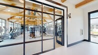 Located at 206 Mountain Creek Dr in Madison, Alabama, this single-story fitness facility offers an expansive, thoughtfully designed space for health and wellness. The architecture immediately impresses with soaring vaulted ceilings supported by prominent exposed natural wood trusses, creating a warm yet dramatic atmosphere throughout the main workout area. Large, black-framed windows line the walls, flooding the interior with abundant natural light and offering views of the surrounding outdoor greenery and pool area.

The main gym floor is generously proportioned, accommodating a comprehensive array of equipment including treadmills, stationary bikes, ellipticals, cable machines, squat racks, free weights, a rowing machine, and a punching bag station. Full-length mirrors and dark accent walls enhance the functional aesthetic of the strength and conditioning zones.

A separate studio room features hardwood flooring, a bold patterned accent wall, a wall-mounted television, and a retractable glass garage door that opens to the outdoors — ideal for group fitness classes or flexible training sessions. The entry corridor is clean and modern, with white tile flooring, decorative sconce lighting, exposed beam accents, and steel-framed glass partitions that provide visual connectivity throughout the facility.